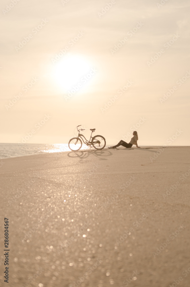 Fototapeta premium Beautiful blonde woman and bicycle traveling down a quiet beach at sunset.