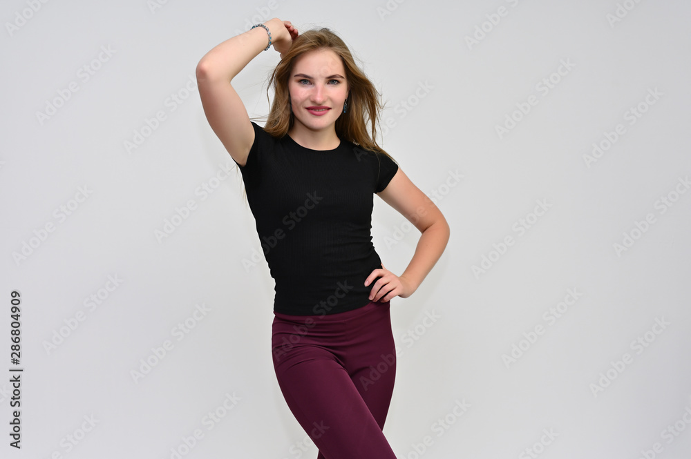 A knee-length portrait of a pretty brunette girl with long flowing hair in a black T-shirt and burgundy trousers on a white background. Smiling, showing emotions. A knee-length portrait of a pretty brunette girl with long flowing hair in a black T-shirt and burgundy trousers on a white background. Smiling, showing emotions.