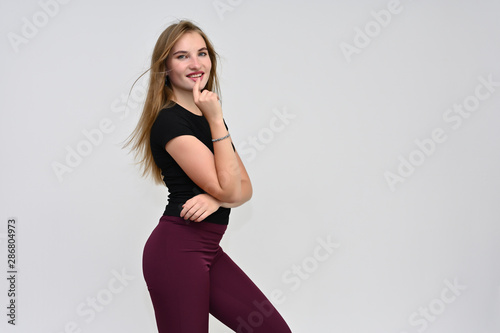 A knee-length portrait of a pretty brunette girl with long flowing hair in a black T-shirt and burgundy trousers on a white background. Smiling, showing emotions.