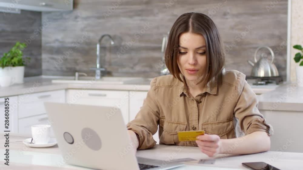 Beautiful caucasian woman sitting at kitchen table and typing credit card information on laptop while buying something online at home Beautiful caucasian woman sitting at kitchen table and typing credit card information on laptop while buying something online at home