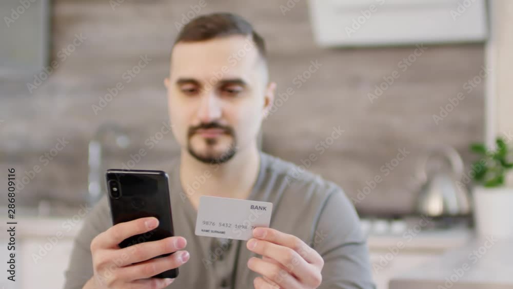 Bearded caucasian man sitting at kitchen table and typing credit card number on smartphone while doing online shopping at home; focus in the foreground Bearded caucasian man sitting at kitchen table and typing credit card number on smartphone while doing online shopping at home; focus in the foreground