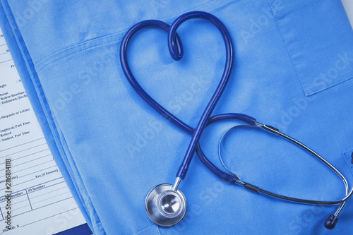 A medical stethoscope is intertwined in the shape of a heart and lies on a medical history and a blue uniform. Closeup of a doctor's uniform.