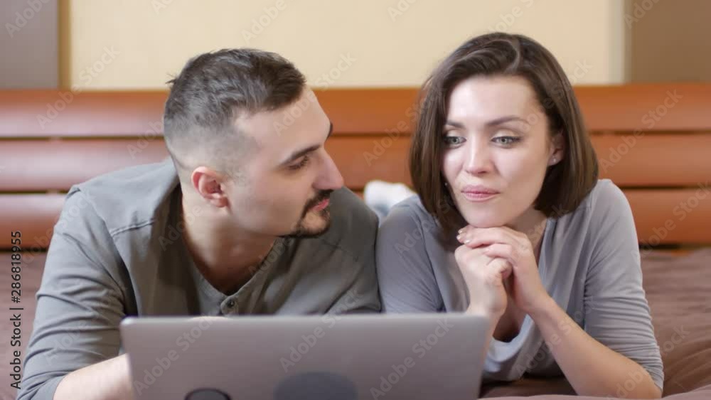 Cheerful couple lying on bed and using credit card and laptop while buying something in online store. Woman happily smiling and clapping hands while husband making payment Cheerful couple lying on bed and using credit card and laptop while buying something in online store. Woman happily smiling and clapping hands while husband making payment