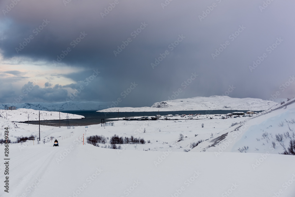Snow covered winter road lead to a small fishing village on the sea in the far North, above the Arctic Circle in Russia's Kola Peninsula. Small snowmobile on the road. Snow covered winter road lead to a small fishing village on the sea in the far North, above the Arctic Circle in Russia's Kola Peninsula. Small snowmobile on the road.