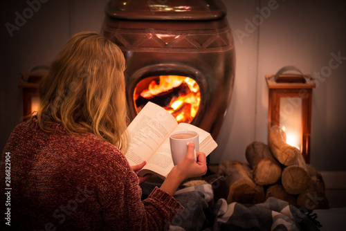 Woman/girl sitting in front of a cozy fireplace during winter under a blanket  reading a book drinking coffee/hot chocolate.