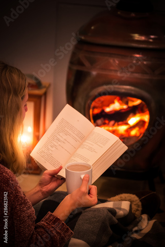 Woman/girl sitting in front of a cozy fireplace during winter under a blanket  reading a book drinking coffee/hot chocolate.