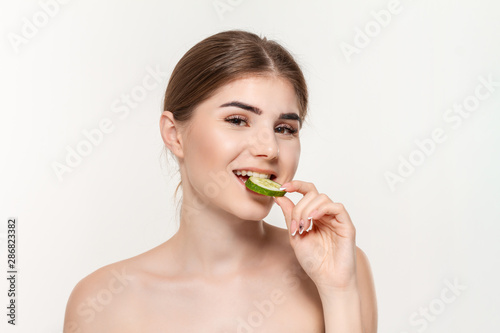 Close-up portrait of a beautiful young girl biting slice of green cucumber solated over white background.