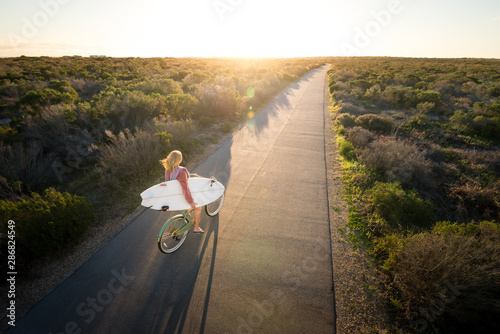 Beautiful blonde surfer girl on her way to the beach on her bicycle with her surfboard.