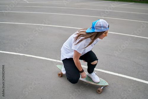 Photo on side of young brunette woman in baseball cap riding skateboard outdoors