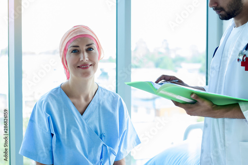 Senior Caucasian woman cover head with a cloth caused of chemotherapy cancer, smiling and sitting on bed, hope to get well. The nurse or carer take care a patient in hospital or health center.