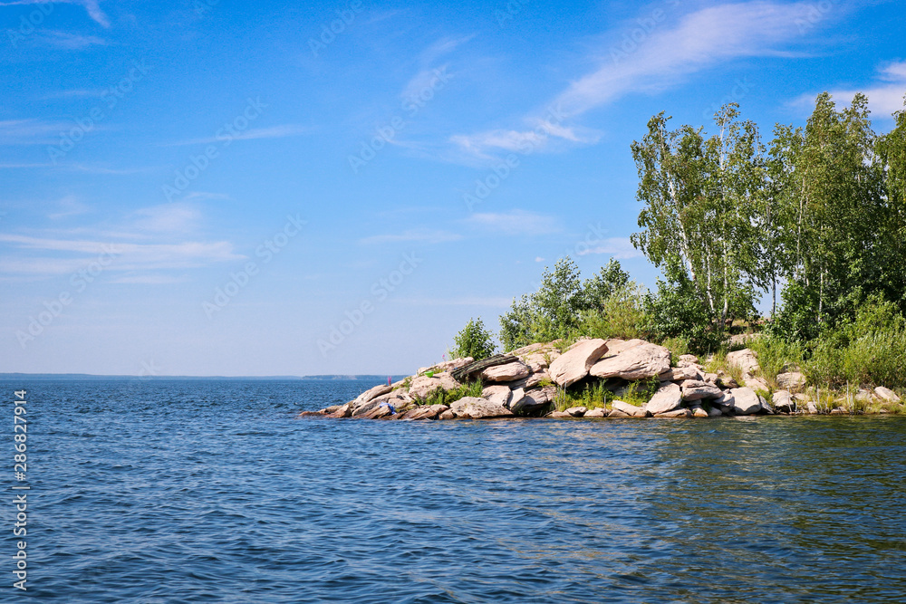 The surface of the lake is covered with ripples and waves against a rocky coast. Summer landscape in sunny weather. The surface of the lake is covered with ripples and waves against a rocky coast. Summer landscape in sunny weather.
