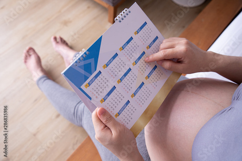 Young pregnant woman counting days with calendar to baby birth on bed in bedroom.