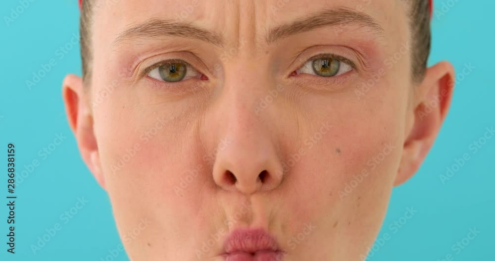 Woman posing at blank studio wall, frowning eyebrows, her look and grimace expressing anger, annoyance and irritation on blue background