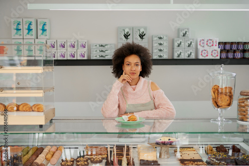 Attractive mixed race pastry shop female worker standing next to counter and looking at camera.