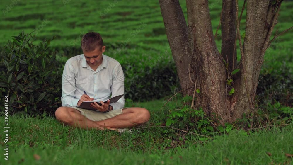 Portrait of a young man sitting under a tree on the grass with a notebook. A man under a tree writes in a notebook, tea plantations in the background. Portrait of a young man sitting under a tree on the grass with a notebook. A man under a tree writes in a notebook, tea plantations in the background.