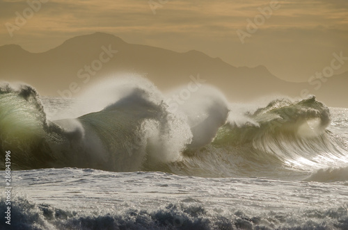 Large wedging waves hitting the coastline under soft sunset light.