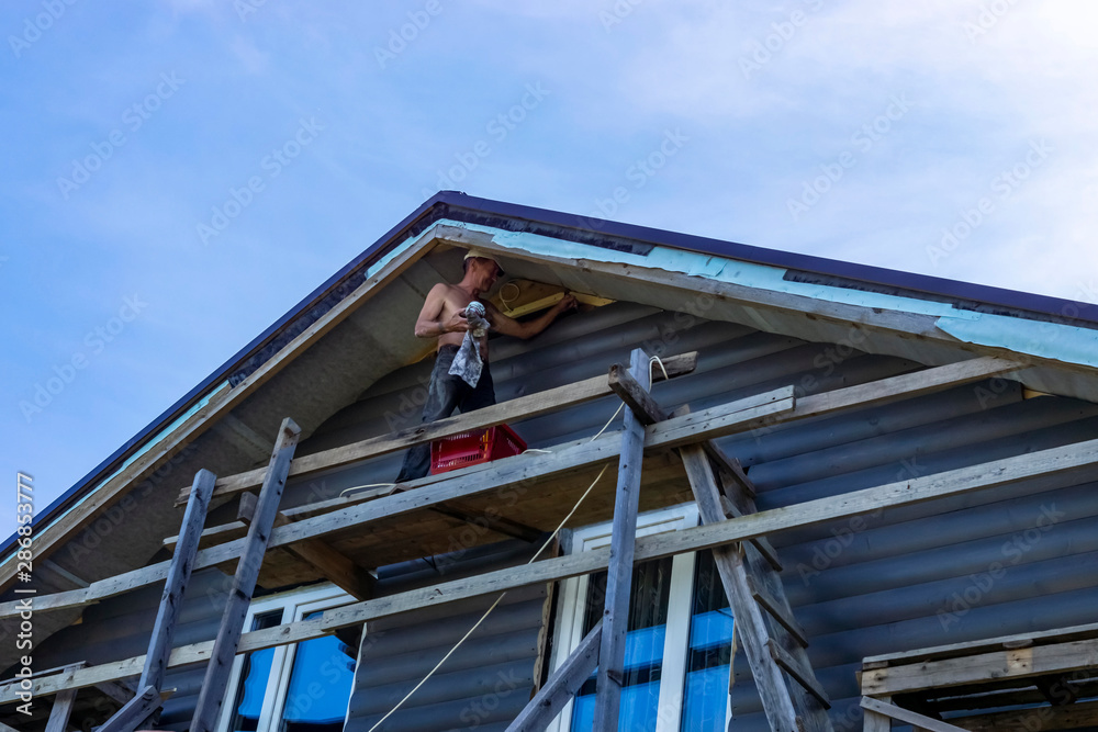 Builder under the roof is engaged in cladding a wooden house wall Builder under the roof is engaged in cladding a wooden house wall