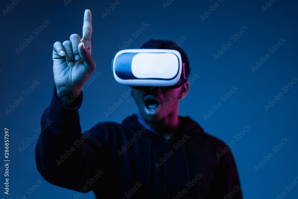 Young african-american man in VR-glasses in neon light on blue background. Male portrait. Concept of human emotions, facial expression, modern gadgets and technologies. Touching empty search bar. Young african-american man in VR-glasses in neon light on blue background. Male portrait. Concept of human emotions, facial expression, modern gadgets and technologies. Touching empty search bar.