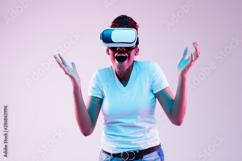 Portrait of young african-american woman's playing in VR-glasses in neon light on gradient background. Concept of human emotions, facial expression, modern gadgets and technologies. Look astonished. © master1305