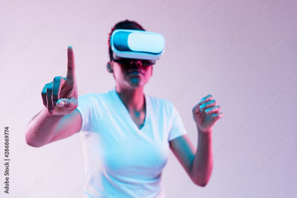 Portrait of young african-american woman's playing in VR-glasses in neon light on gradient background. Concept of human emotions, facial expression, modern gadgets and technologies. Touches empty bar. Portrait of young african-american woman's playing in VR-glasses in neon light on gradient background. Concept of human emotions, facial expression, modern gadgets and technologies. Touches empty bar.