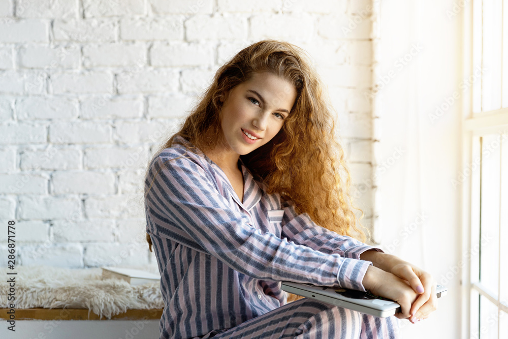 Portrait of good-looking model with luxury curly hair and attractive smile posing on white brick wall and holding trendy notebook. Morning pyjamas and modern interior concept
