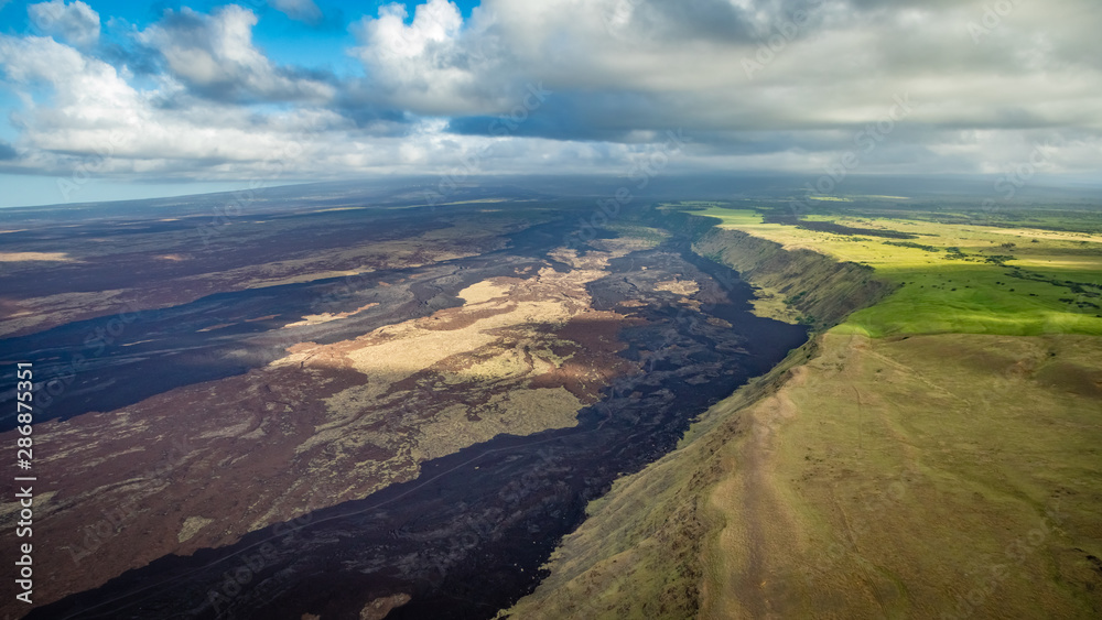 Aerial view of green fields on the right with a steep ridge leading down to lava fields over Kaimu'Uala on the left