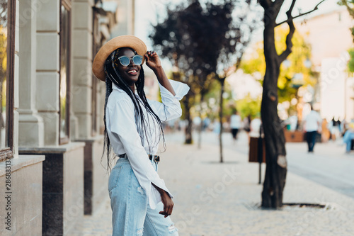 Portrait of a smiling young African American girl with pigtails in a fashionable hat and stylish sunglasses walking along the street on a sunny day