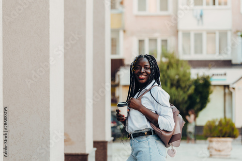 Portrait of a smiling young African American girl with pigtails with coffee walking in the street on a sunny day. Outdoor photo. © Nana_studio