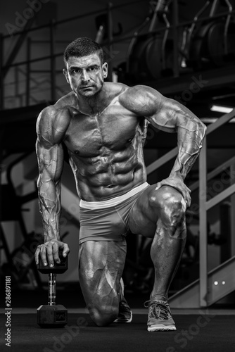 Bodybuilder Posing in the Gym, Kneeling on the Floor With his hand on Dumbbell. Black and White Image