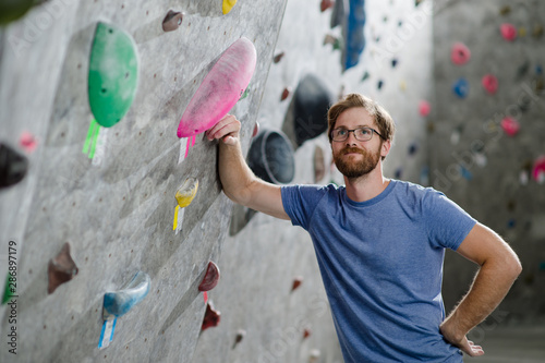 cute happy active sport caucasian man with beard wearing glasses leaning on climbing wall indoors
