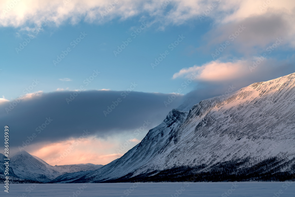 Fototapeta premium Snow covered valley with Rahpattjarro Lapland Sweden during calm morning with warm sunbeam, dark band of clouds, blue sky