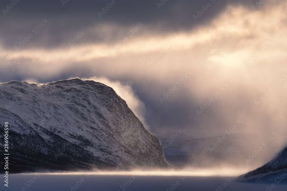 Dramatic nordic winter landscape in Lapland Sweden during snow storm with mystic light, sun, mist, and dark cloud layer