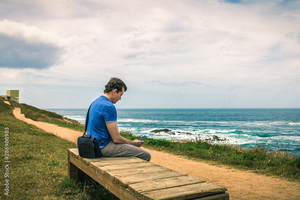 man sitting on a bench using smart phone facing the sea next to the pathway