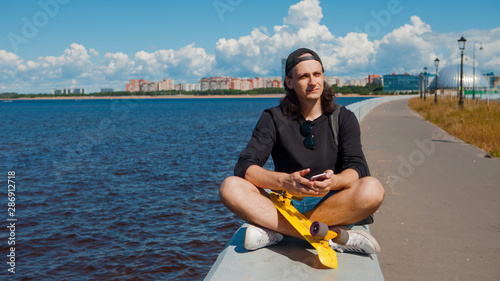 A young man sitting on a quay with a yellow skateboard on his lap is checking a smartphone.