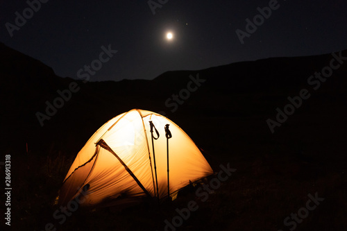 The camping tent is lit by a lantern at night in the mountains. Bright moon in the sky. Mountain climbing hike.