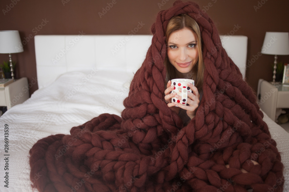 cute young woman is sitting on the bed wrapped in a big and fluffy brown plaid. Beautiful girl in a cozy blanket - obrazy, fototapety, plakaty 