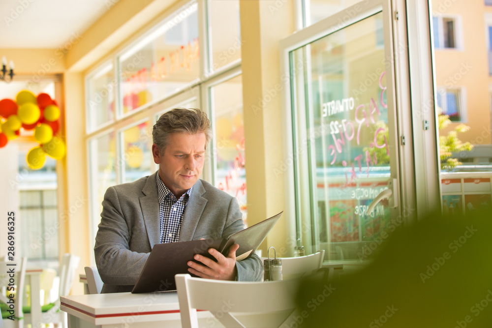 Mature customer reading menu while sitting at table by window in restaurant