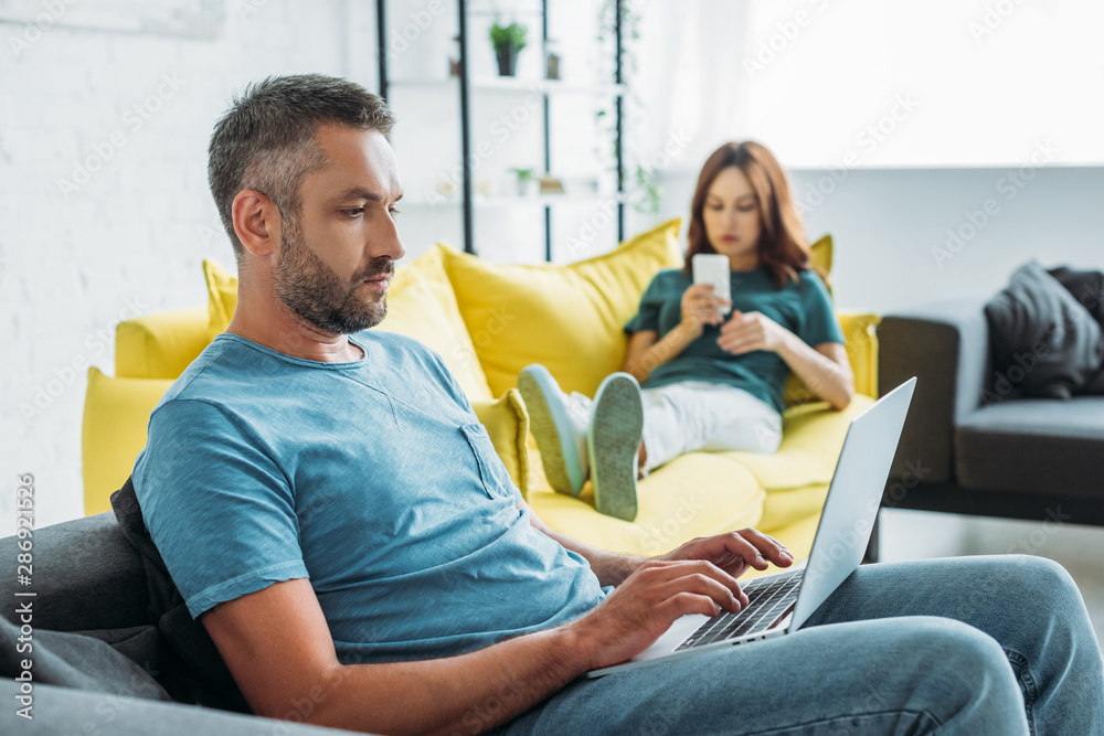 selective focus of serious man using laptop while sitting on sofa near wife using smartphone selective focus of serious man using laptop while sitting on sofa near wife using smartphone