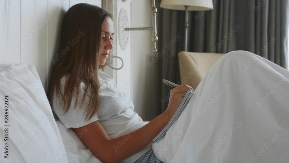 Young woman sitting on bed and writing morning pages to her diary after awake in bedroom. Female writing notes in domestic interior. Young woman sitting on bed and writing morning pages to her diary after awake in bedroom. Female writing notes in domestic interior.