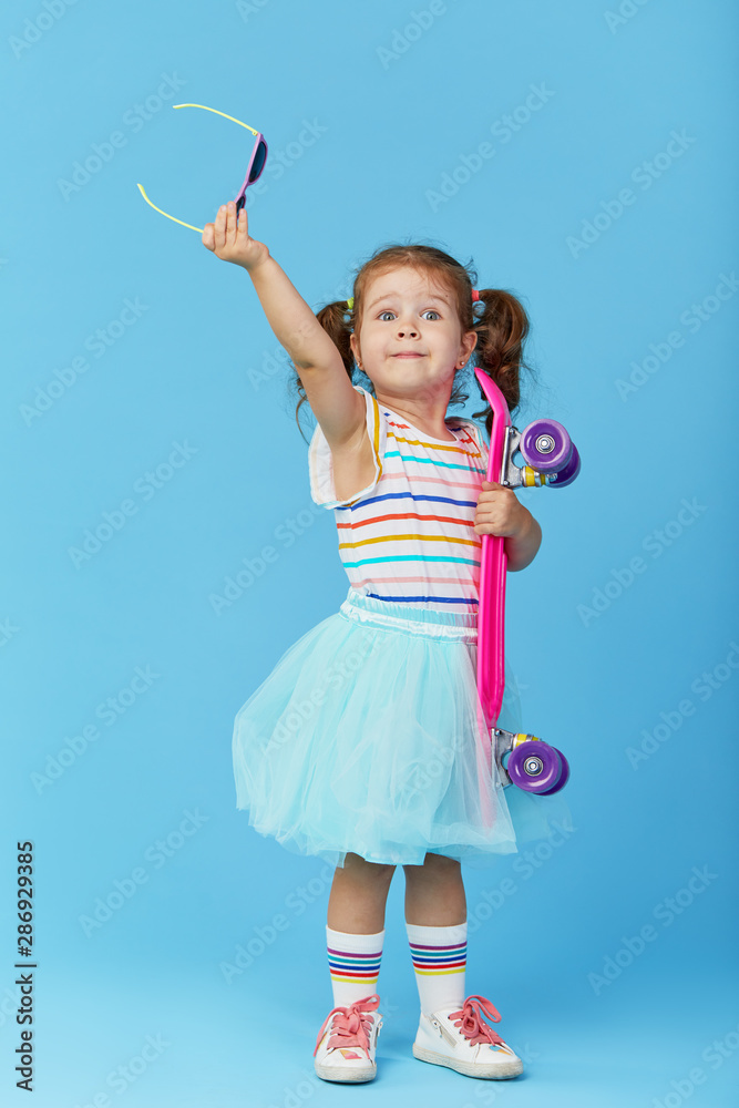 Stylish cool little girl child in bright clothes with skateboard over blue background. Space for text Stylish cool little girl child in bright clothes with skateboard over blue background. Space for text
