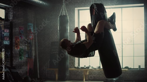 Insane Punching Bag Crunches. Strong Masculine Man Embrassing the Bag with His Legs and Pumping His Abs and Core Muscles. Handsome Male Trains in a Gym with Motivational Posters. photo