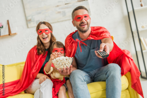 selective focus of father holding remote controller while family in superheroes costumes sitting on sofa with popcorn and watching tv