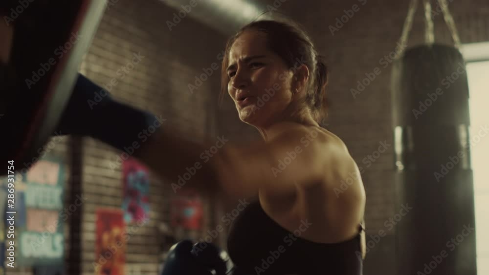 Fit Athletic Woman Kickboxer Punches and Hits the Punching Pads During a Workout in a Loft Gym. She's Beautiful and Energetic. Strong Coach is Holding the Boxing Pads. Intense Self-Defence Training. Fit Athletic Woman Kickboxer Punches and Hits the Punching Pads During a Workout in a Loft Gym. She's Beautiful and Energetic. Strong Coach is Holding the Boxing Pads. Intense Self-Defence Training.