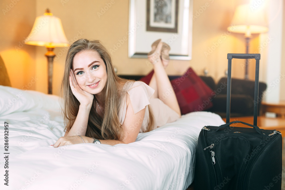 Smiling young businesswoman in bed staring at camera. Hotel room. the girl licks on the bed near the suitcase.