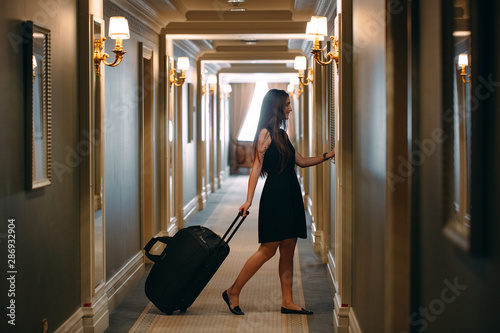 Young woman with handbag and suitcase in an elegant suit walks the hotel corridor to her room.
