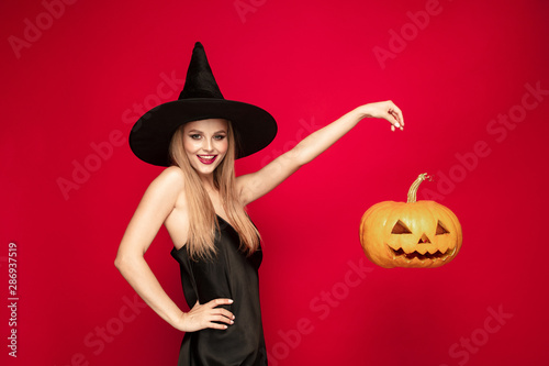 Young blonde woman in black hat and costume on red background. Attractive caucasian female model. Halloween  black friday  cyber monday  sales  autumn concept. Copyspace. Flying pumpkin near.