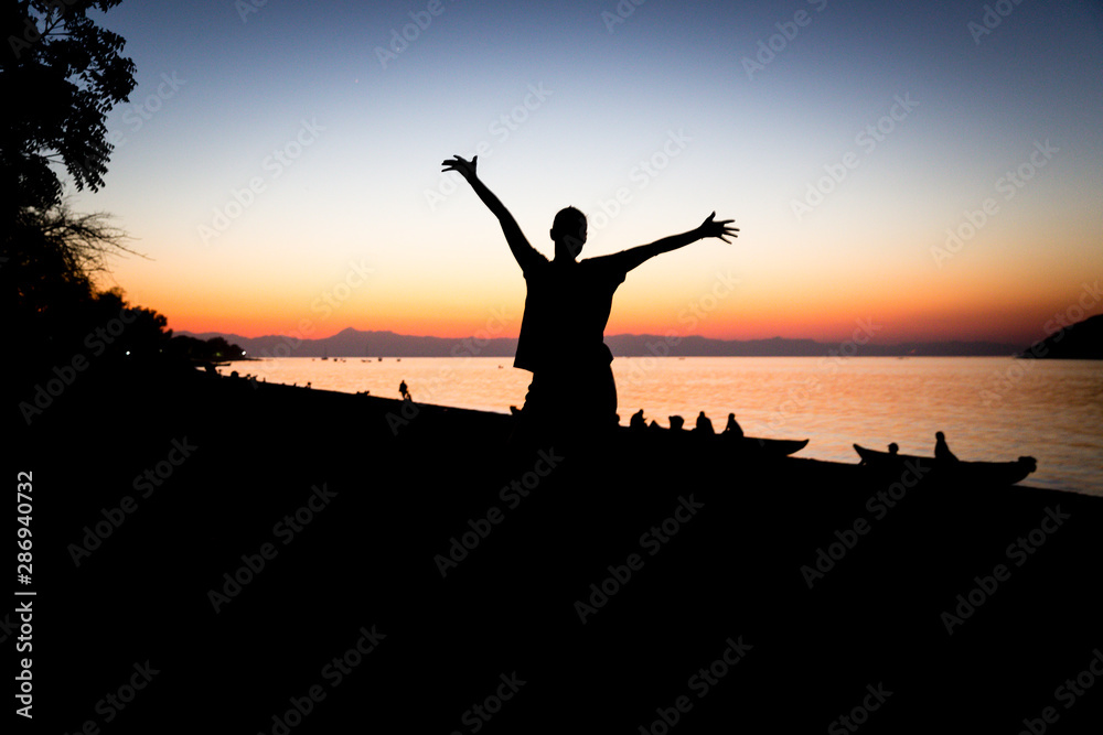 Lake Malawi at Monkey Bay, Woman silhouette is raising arms, people in the background gathering togehter at the Beach, washing dishes, talking, red sunset, South-East-Africa