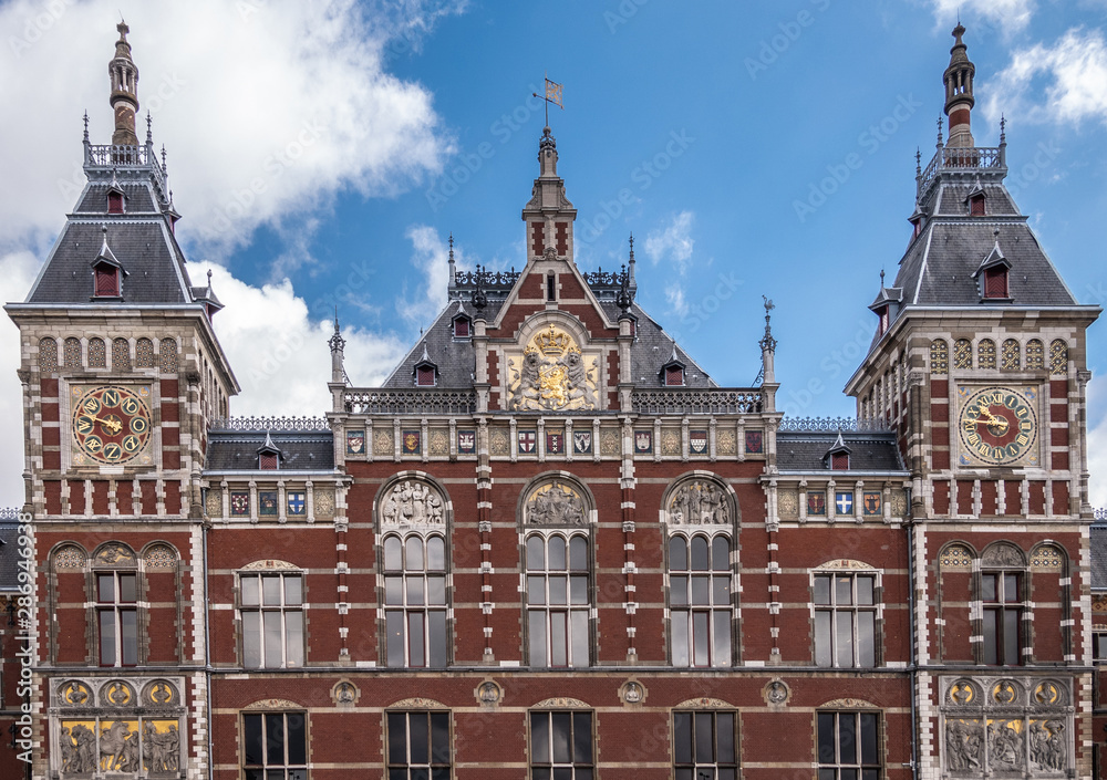 Red brick with white trim upper part facade Centraal Railway Station with towers, clocks, and plenty of frescoes under blue sky with white clouds.