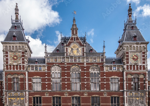 Red brick with white trim upper part facade Centraal Railway Station with towers, clocks, and plenty of frescoes under blue sky with white clouds. © Klodien