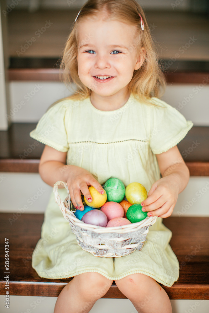 Fototapeta premium Cute little child girl with blonde hair on Easter day. Girl holding basket with painted eggs and sitting on the stairs at home